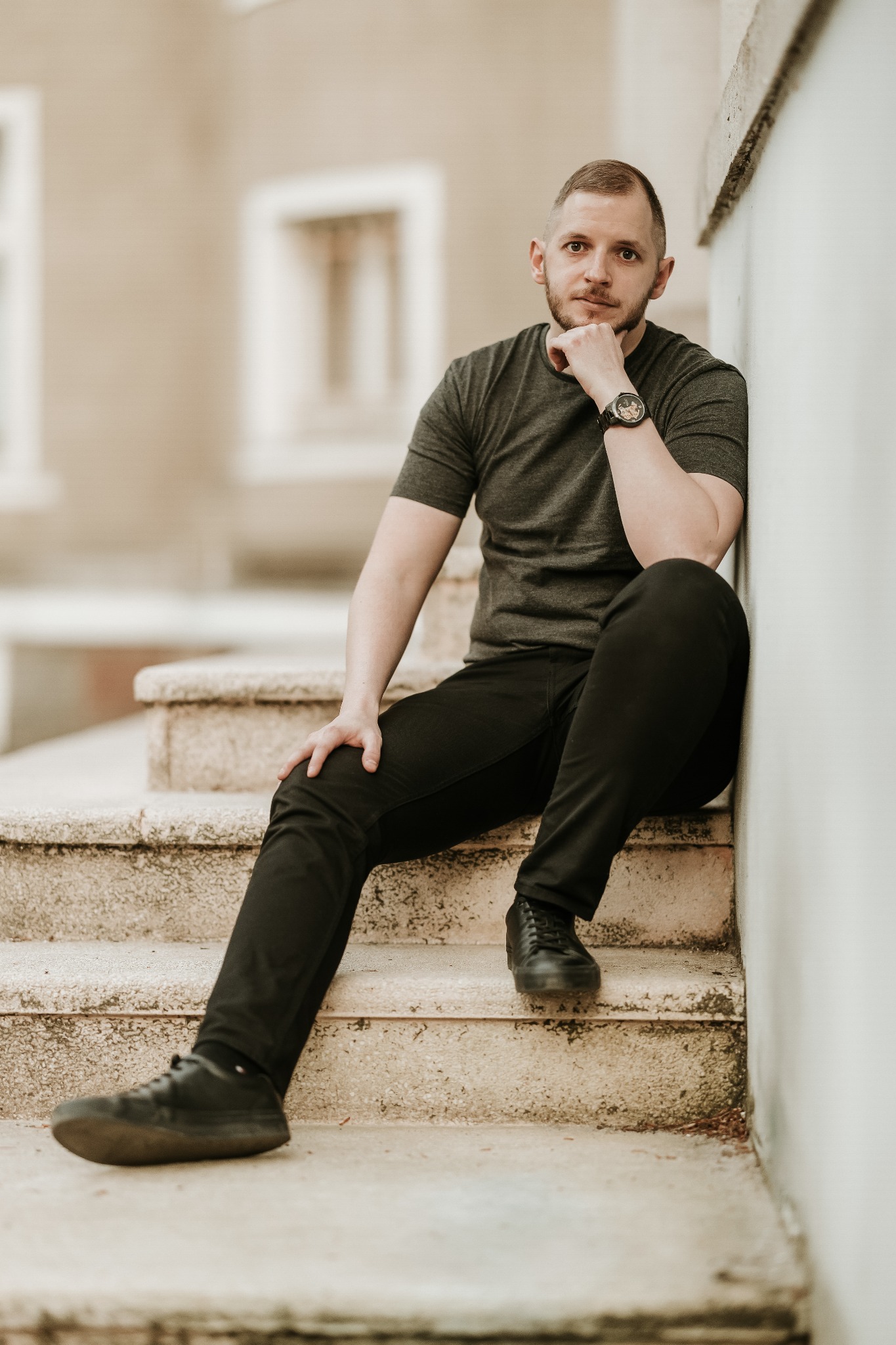 Mężczyzna z brodą siedzi na kamiennych schodach, oparty o ścianę, z ręką podpierającą brodę i zegarkiem na nadgarstku.