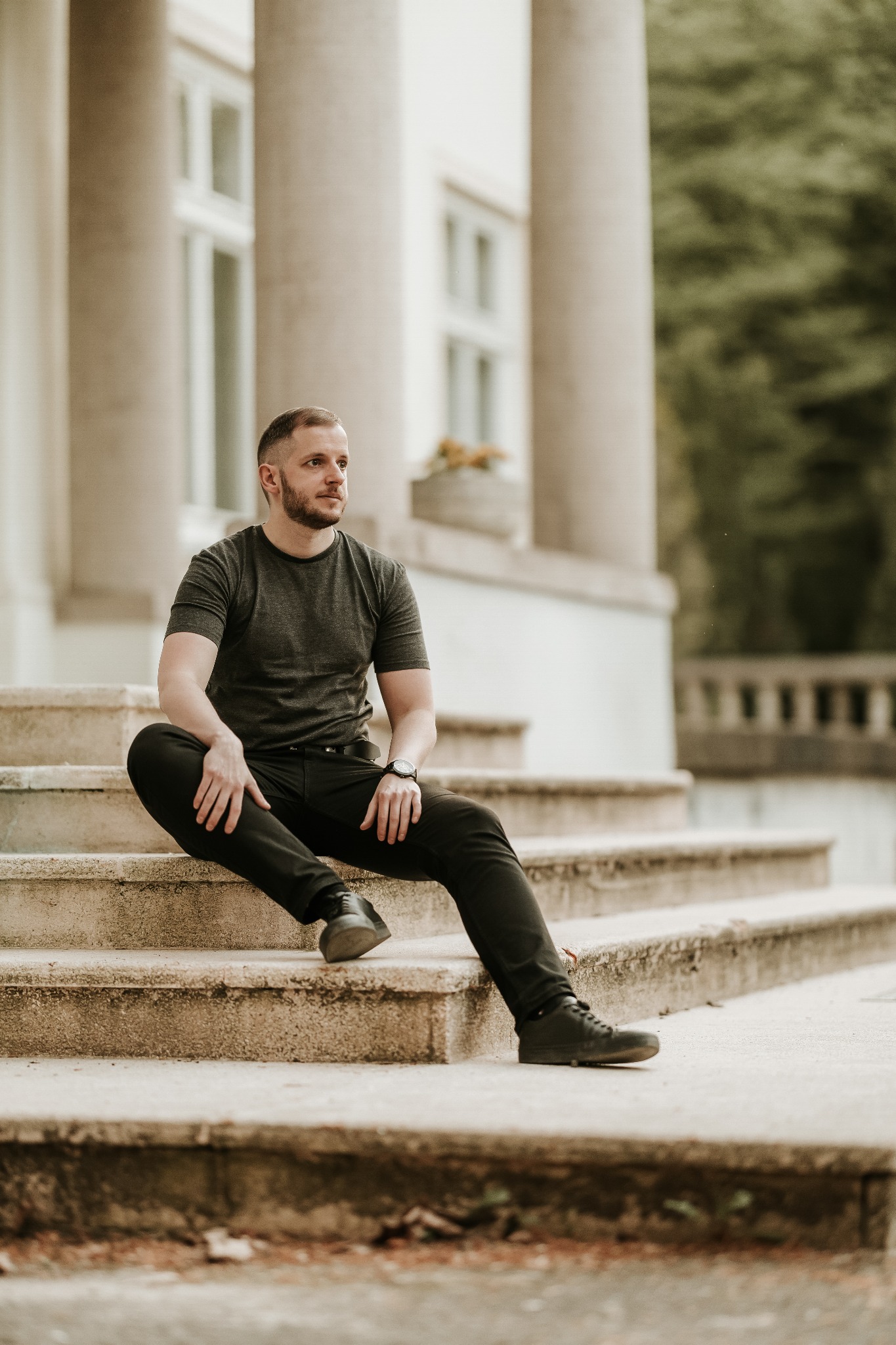 Mężczyzna w czarnym ubraniu siedzi na kamiennych schodach klasycznego budynku z kolumnami, patrząc w dal.