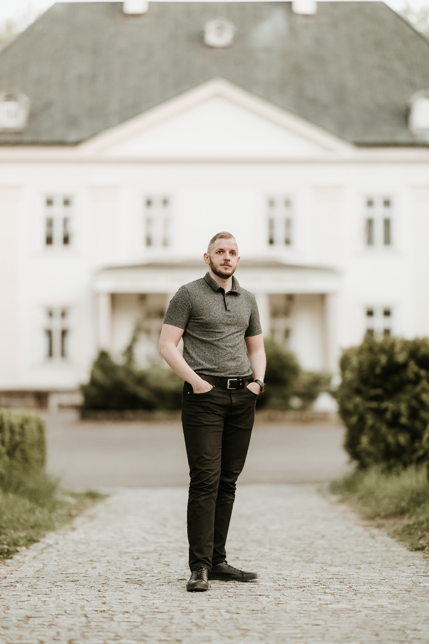 Mężczyzna w szarej koszulce polo i czarnych spodniach pozuje na brukowanej ścieżce przed białym budynkiem z szarym dachem.