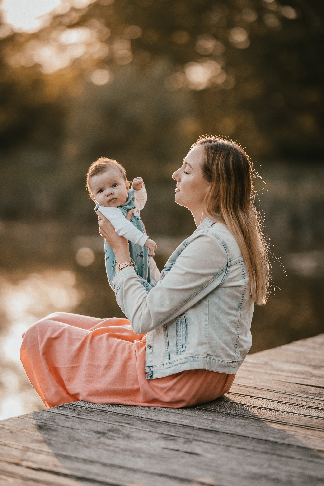 Kobieta siedzi na drewnianym pomoście, trzymając na rękach niemowlę w błękitnym kombinezonie z motywem pojazdów, w tle woda i drzewa w ciepłym świetle.