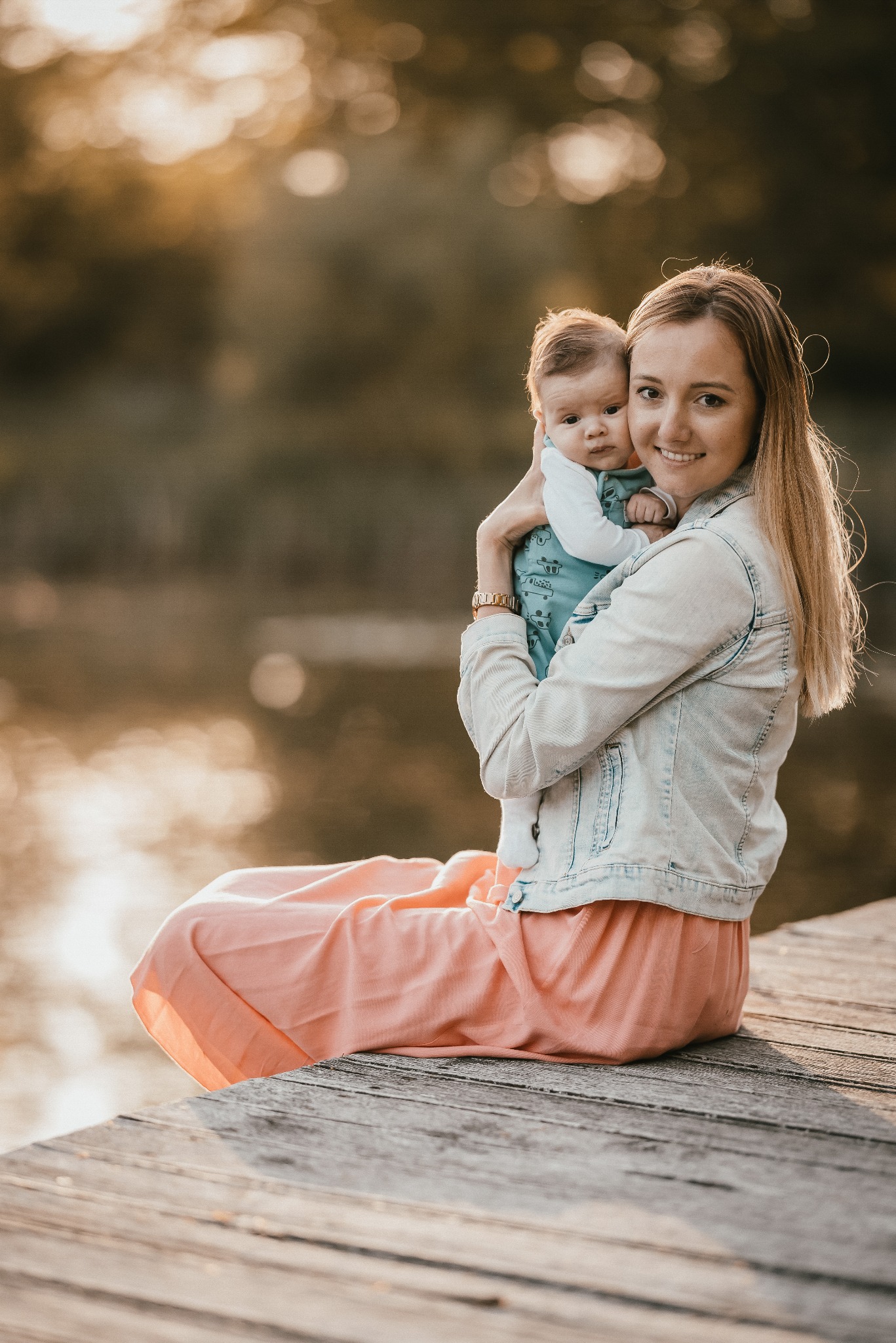 Uśmiechnięta mama w dżinsowej kurtce i łososiowej spódnicy siedzi na drewnianym pomoście, trzymając na rękach niemowlę ubrane w błękitne body z nadrukiem pojazdów; w tle woda i drzewa w ciepłym...