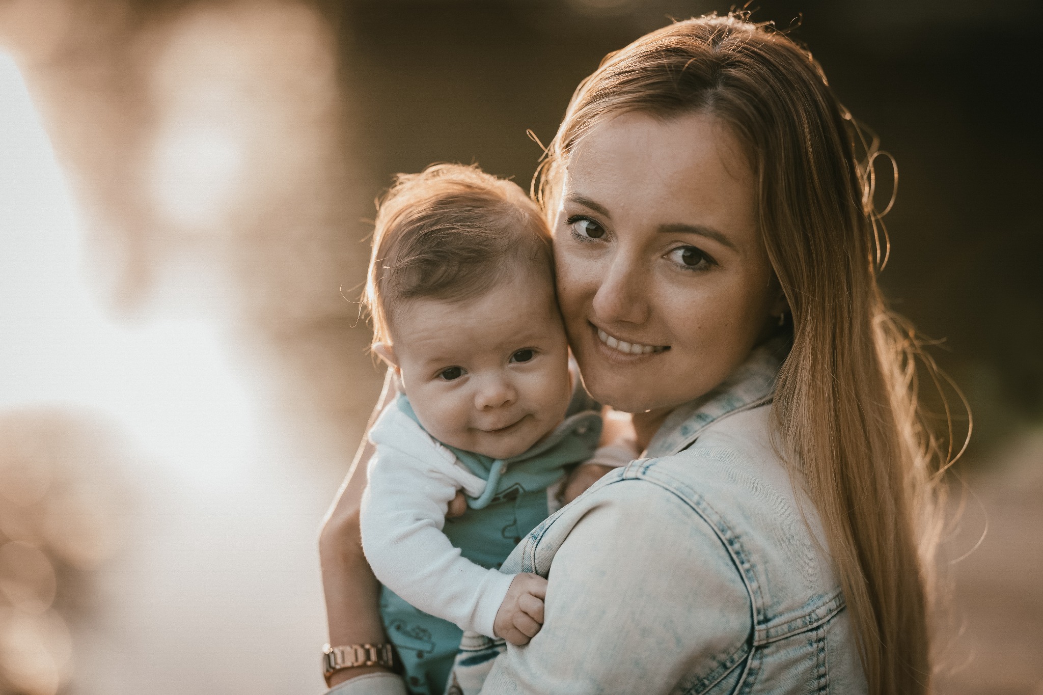 Uśmiechnięta matka trzymająca na rękach niemowlę, oboje patrzą w obiektyw, ciepłe światło słoneczne