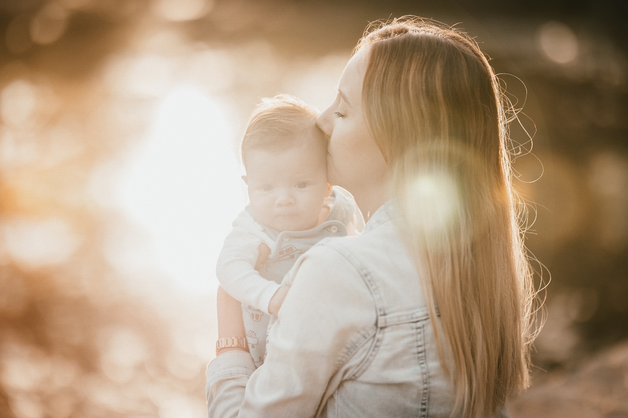 Matka całuje w czoło trzymane na rękach niemowlę ubrane w jasne ubranko, ciepłe światło zachodzącego słońca tworzy miękkie tło.