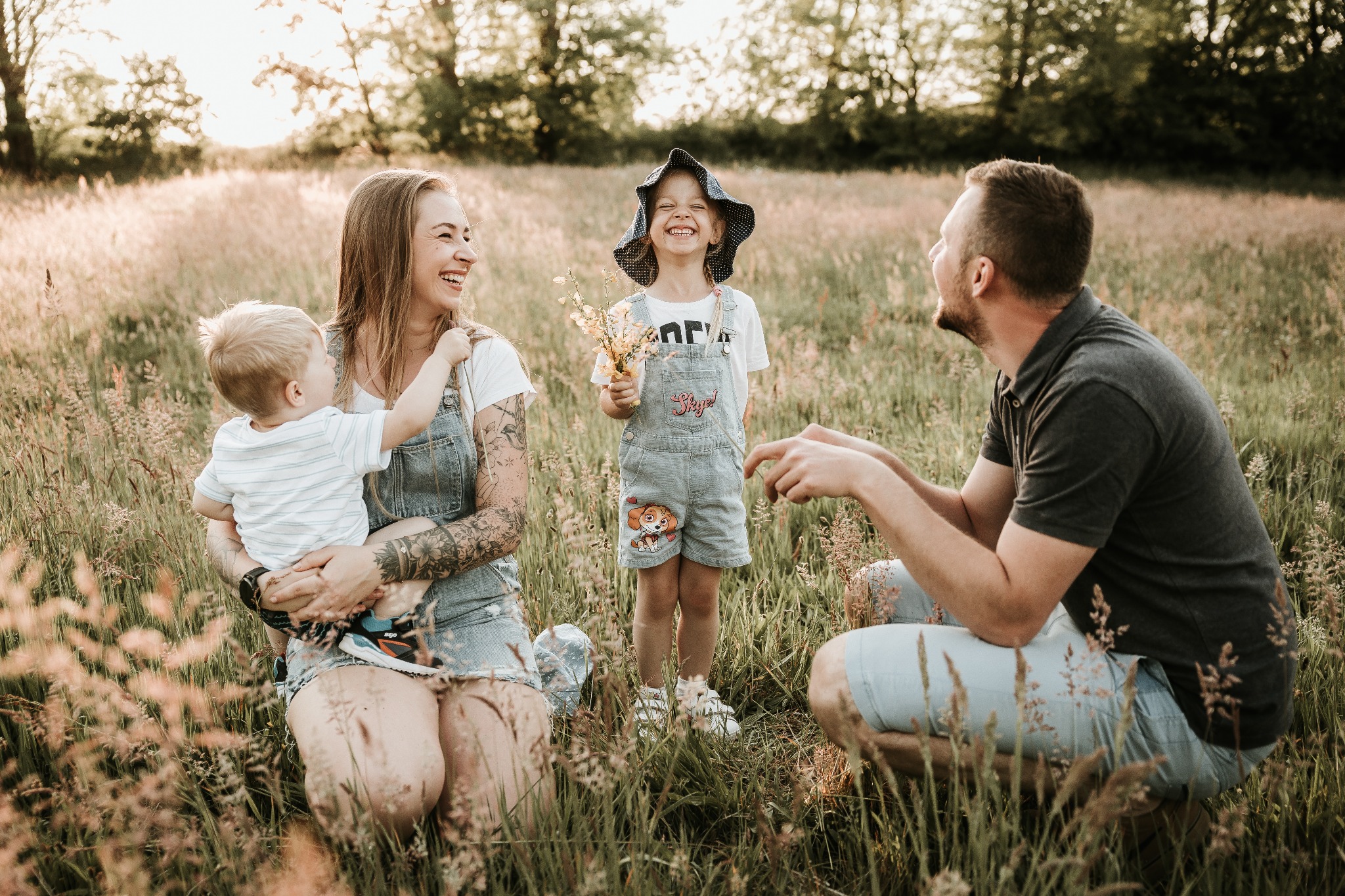 Szczęśliwa rodzina z dwójką dzieci w polu wysokiej trawy, mama trzyma synka na rękach, córka stoi z kwiatami, tata kuca obok.