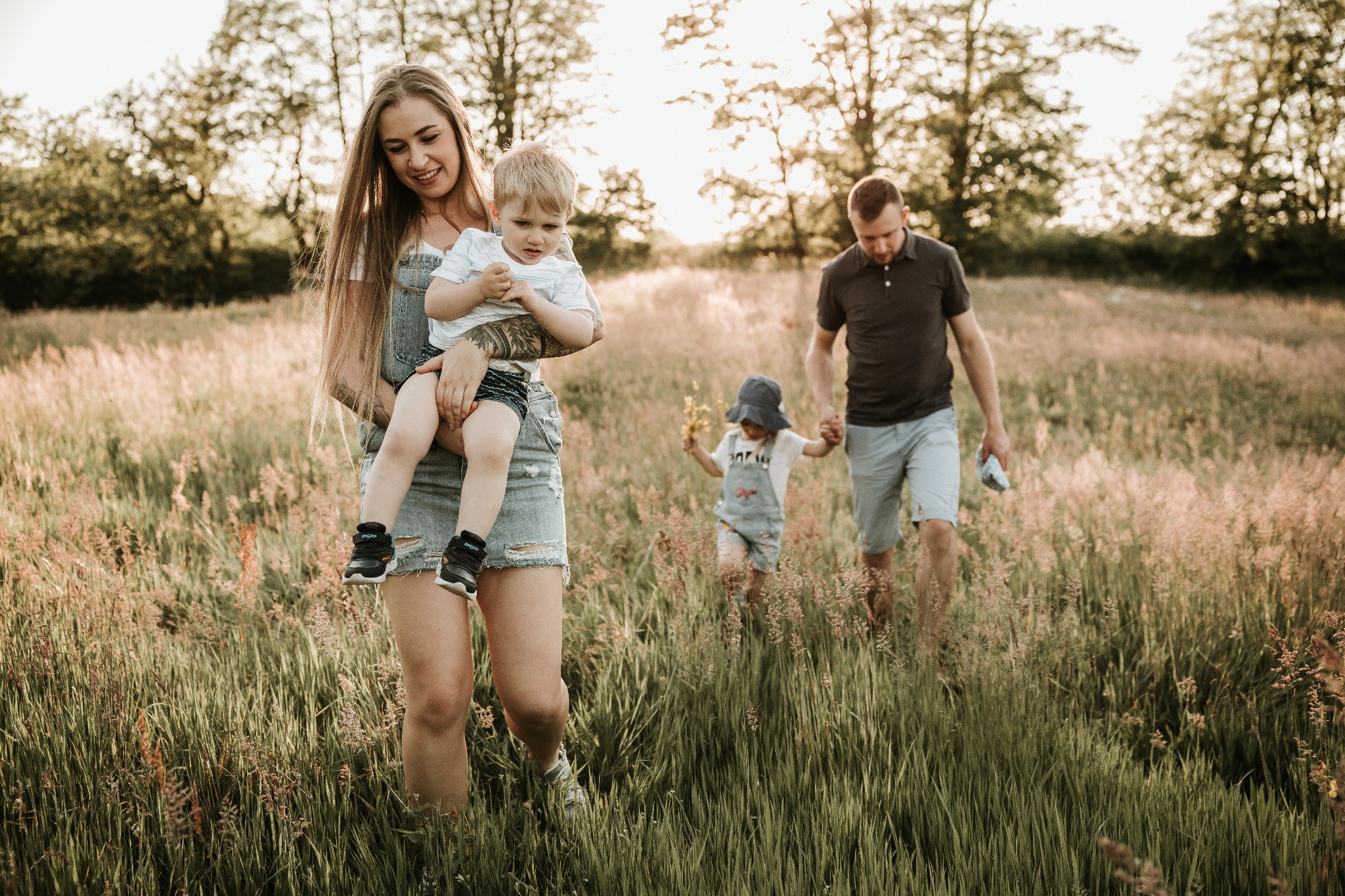 Rodzina na spacerze po łące: mama trzyma synka na rękach, tata i córka idą za nimi, trawa wysoka, delikatne światło zachodzącego słońca.