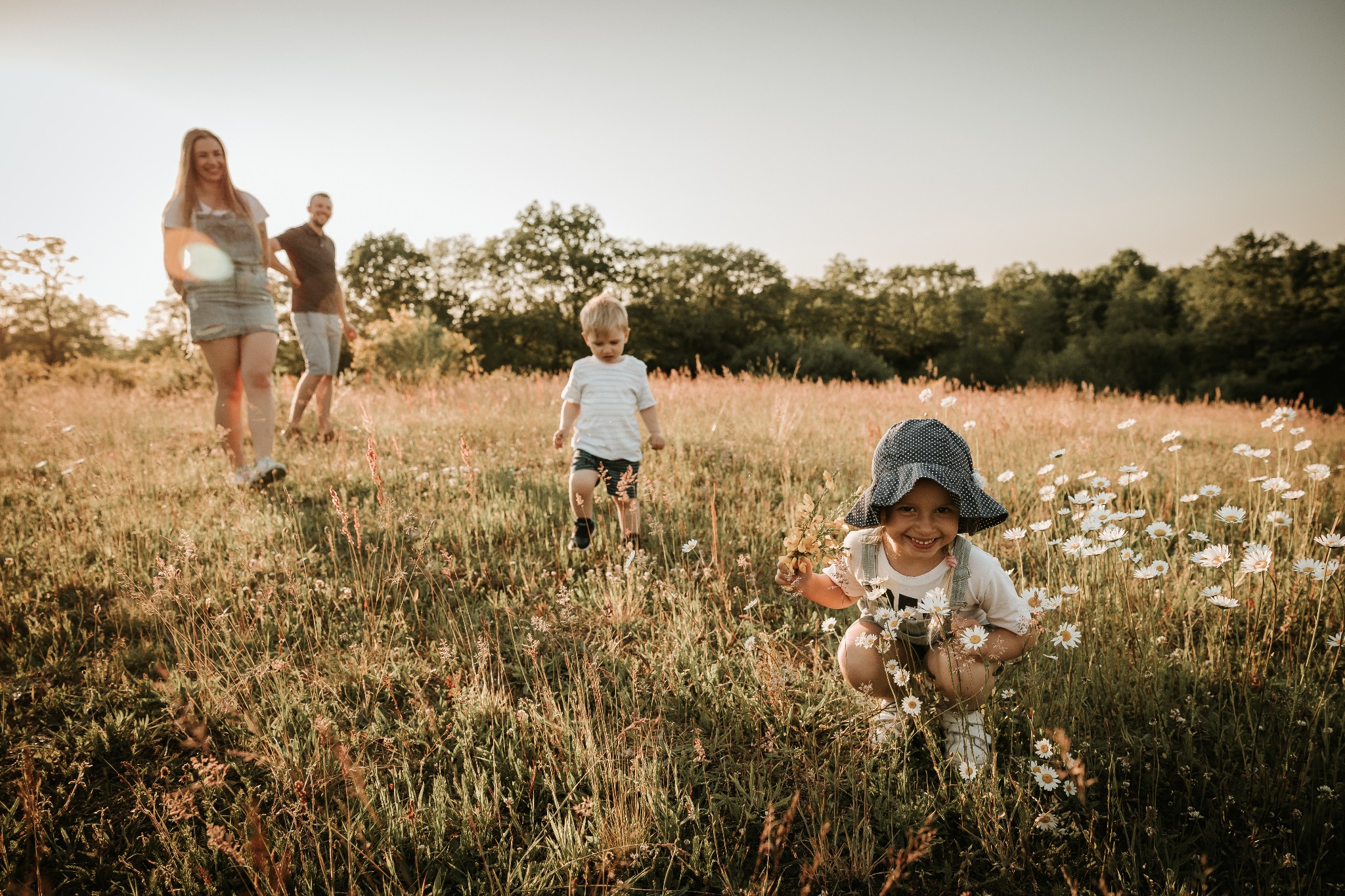 Dziewczynka w kapeluszu kuca w łące pełnej rumianków, trzymając bukiet kwiatów, w tle rodzina spaceruje pośród traw w ciepłym świetle zachodzącego słońca.