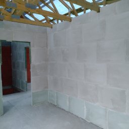 DomoStart Android Povkh - Wnętrze domu w trakcie budowy, widoczne ściany z bloczków, surowy stan, drewniana konstrukcja dachu bez pokrycia, widoczne otwory drzwiowe.