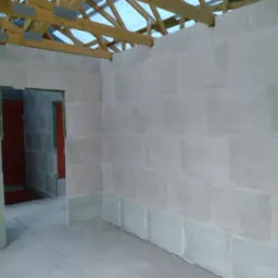 Wnętrze domu w trakcie budowy, widoczne ściany z bloczk&oacute;w, surowy stan, drewniana konstrukcja dachu bez pokrycia, widoczne otwory drzwiowe.