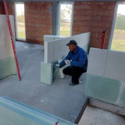 DomoStart Android Povkh - Pracownik w niebieskiej bluzie, w pozycji kucznej, nakłada zaprawę na jasne płyty w surowym wnętrzu budynku w trakcie budowy. Widoczne surowe ściany z cegły, okna bez ram i poziomica oparta o ścianę.