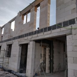 DomoStart Android Povkh - Wznoszenie ścian budynku z betonu komórkowego, widoczne szare bloczki na wysokości stropu, podpory stropu w otworach drzwiowych, siatka zbrojeniowa w spoinach, oraz pracownik w oddali.