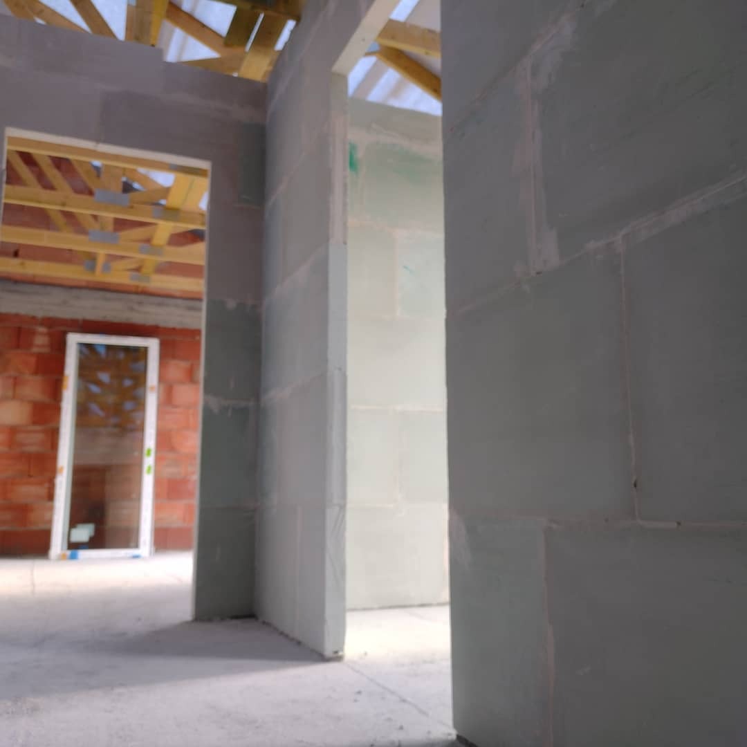 Wnętrze w trakcie budowy: widoczne surowe ściany z betonu komórkowego, drewniana konstrukcja dachu, okno wstawione w ścianę z czerwonej cegły, surowy beton na podłodze.