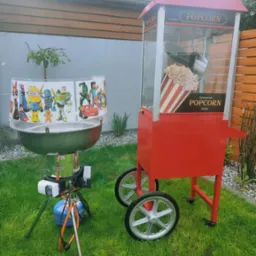 Dwa urządzenia do przygotowywania i serwowania popcornu na trawie: profesjonalny wózek z popcornem w stylu retro z napisem 'Popcorn' oraz przenośny kociołek z osłoną z postaciami z bajek.
