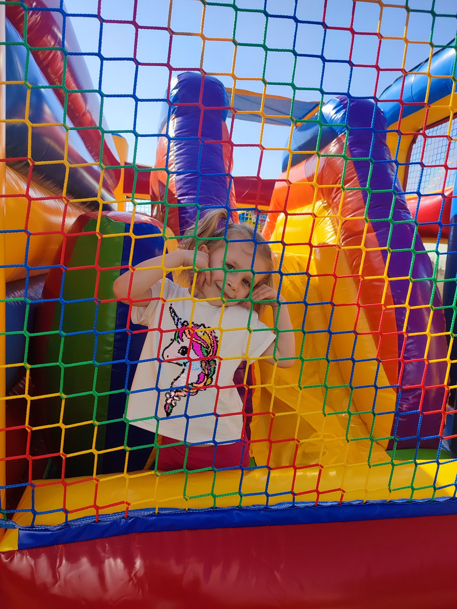 Dziewczynka w koszulce z jednorożcem wygląda przez kolorową siatkę dmuchanego zamku.