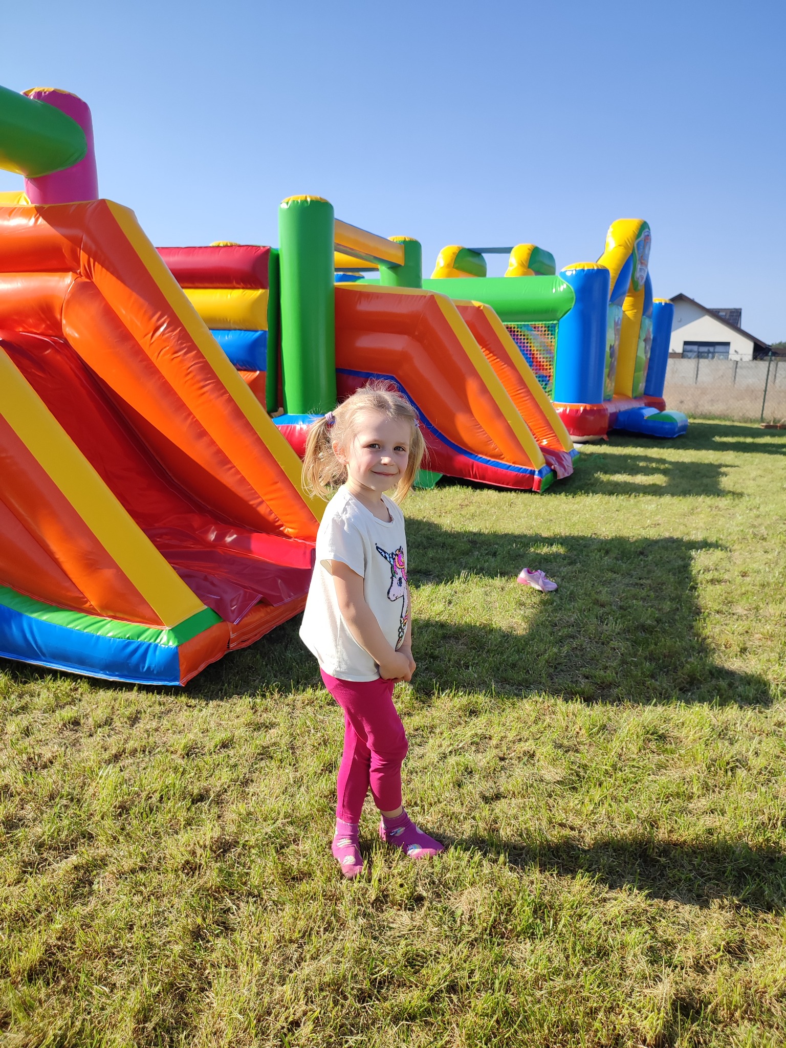 Uśmiechnięta dziewczynka z blond włosami w kucykach, ubrana w różowe legginsy i białą koszulkę z jednorożcem, stoi na trawie przed wielokolorowym, dmuchanym placem zabaw z elementami zjeżdżalni...