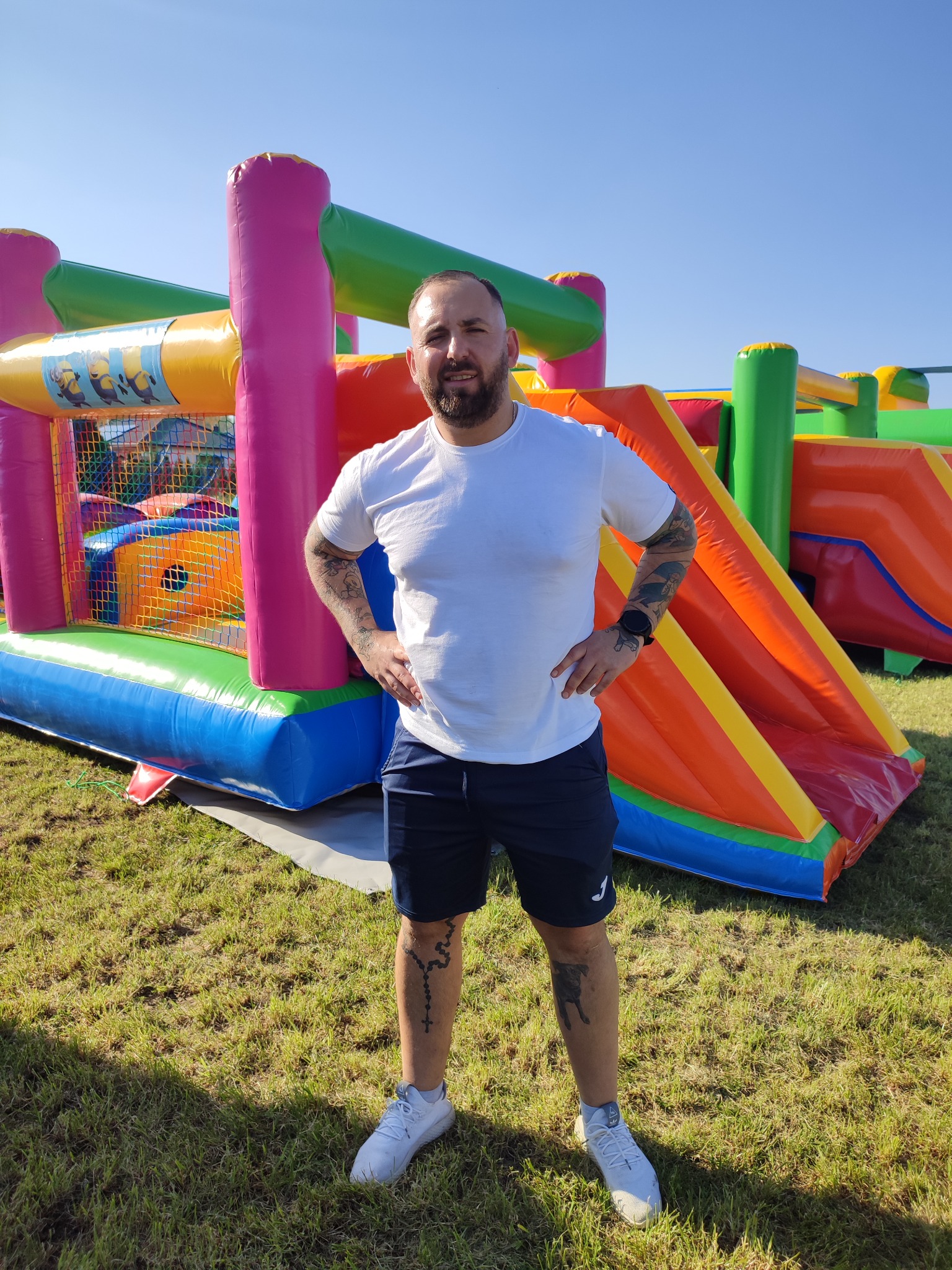 Mężczyzna w białej koszulce i granatowych spodenkach stoi na trawie przed kolorowym dmuchanym zamkiem z motywem Minionków i zjeżdżalniami, z tatuażami na rękach i nogach, pozujący z rękami...