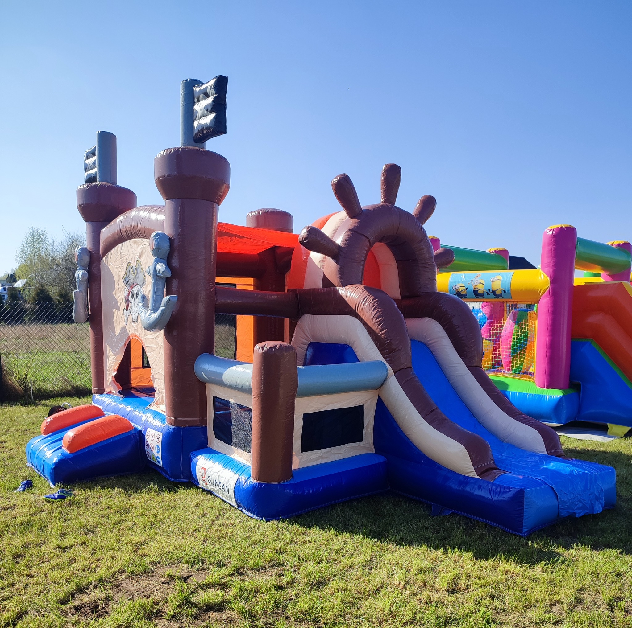 Kolorowy, dmuchany zamek w kształcie statku pirackiego z elementami zjeżdżalni i motywami z bajek, ustawiony na trawie.