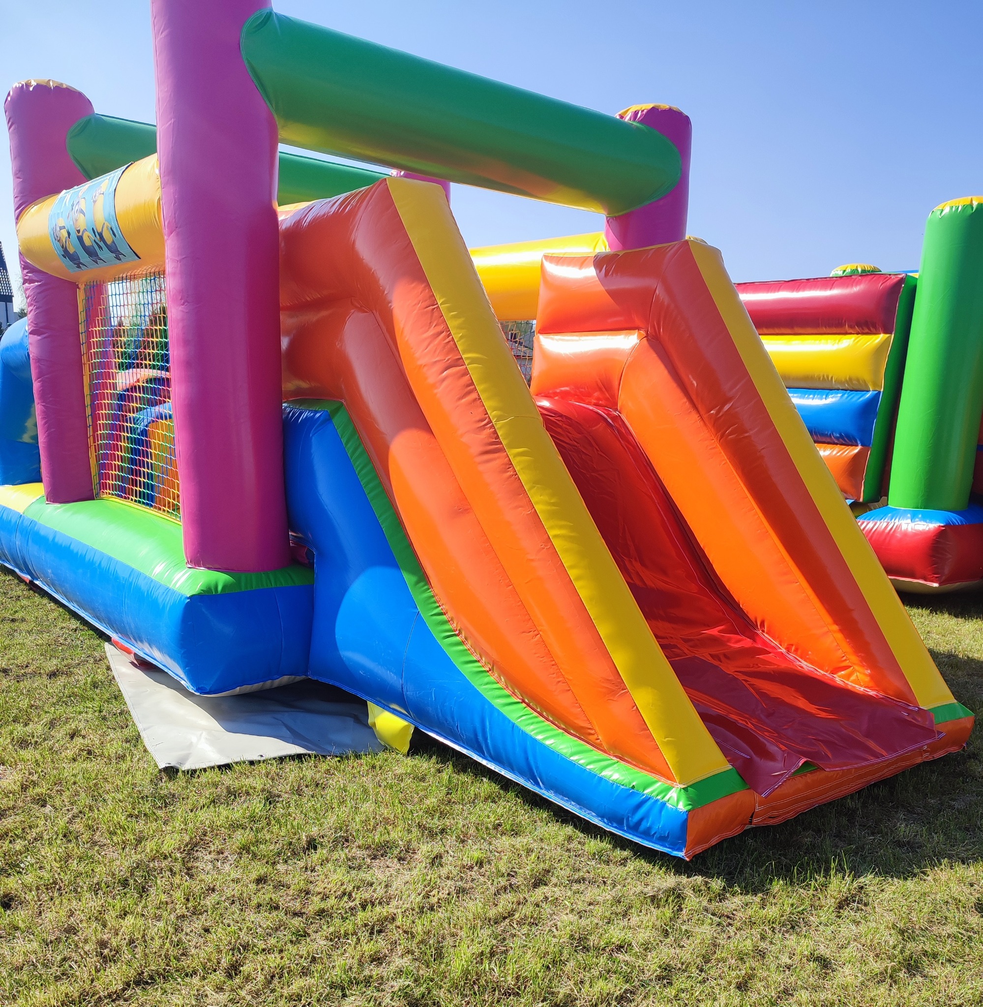 Kolorowy dmuchany zamek ze zjeżdżalnią na trawie, widoczne mocne szwy i detale konstrukcyjne, w tle fragmenty innych dmuchańców, błękitne niebo.