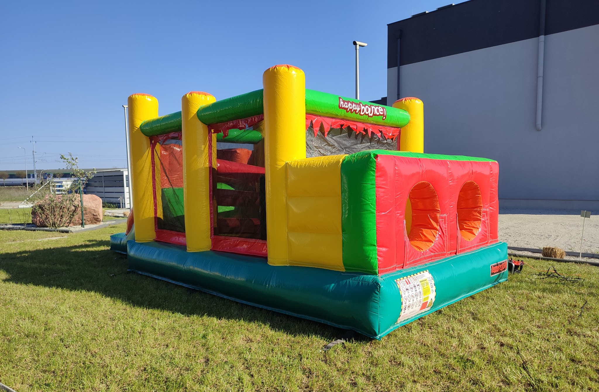 Kolorowy dmuchany zamek z napisem 'happy bounce' na zielonej trawie w słoneczny dzień, z widocznym budynkiem w tle.