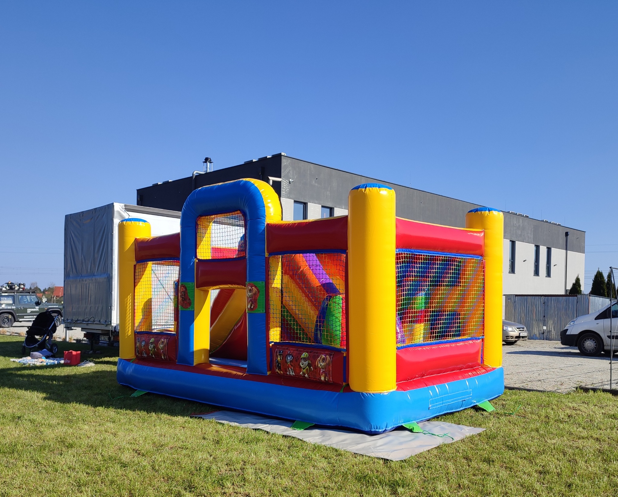 Kolorowy, dmuchany zamek z motywem bajki Psi Patrol ustawiony na trawie przed budynkiem i zaparkowanymi samochodami.
