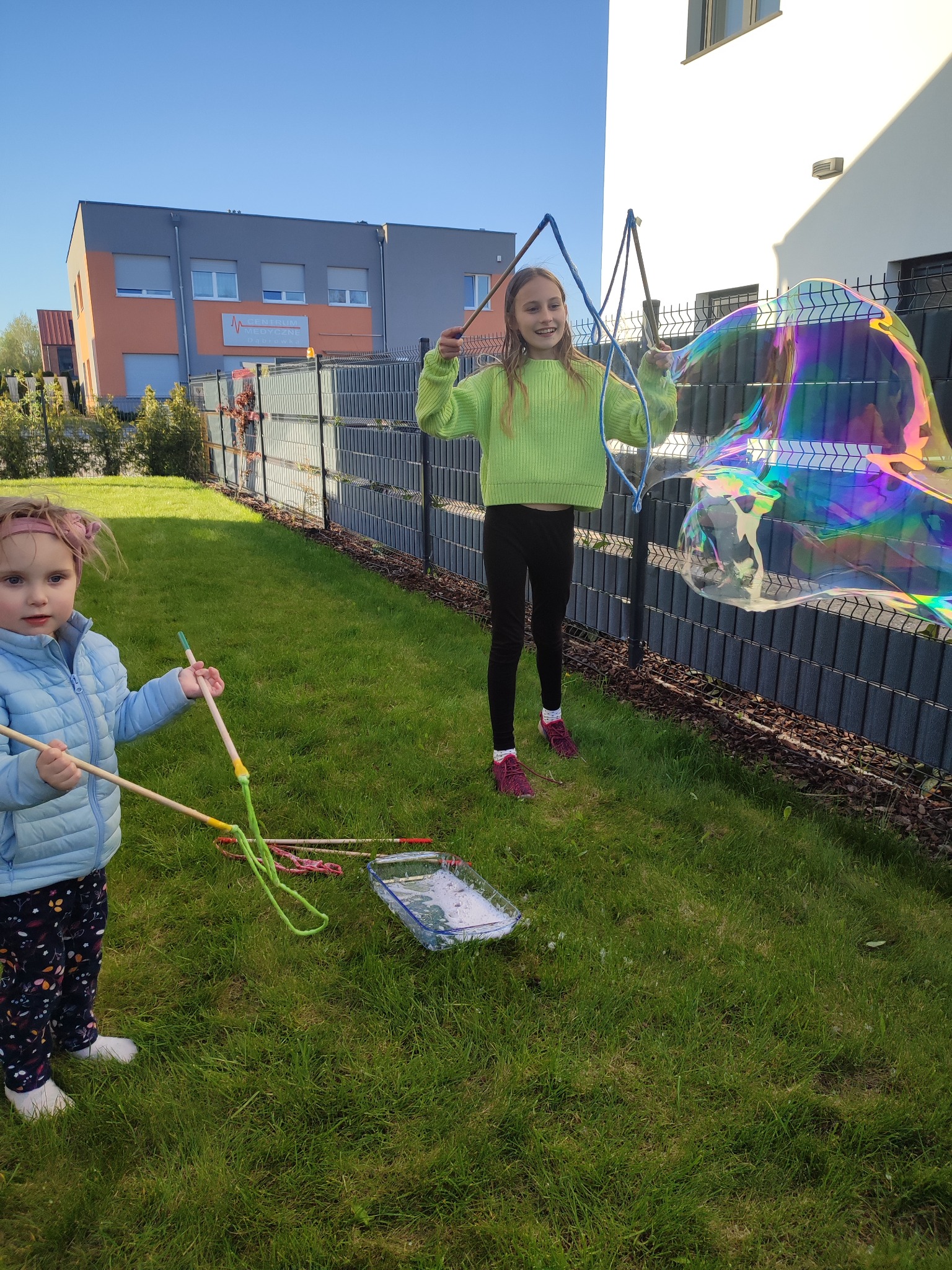 Dwie dziewczynki bawią się w puszczanie dużych baniek mydlanych na trawniku przed domem, używając specjalnych sznurków i kijków.