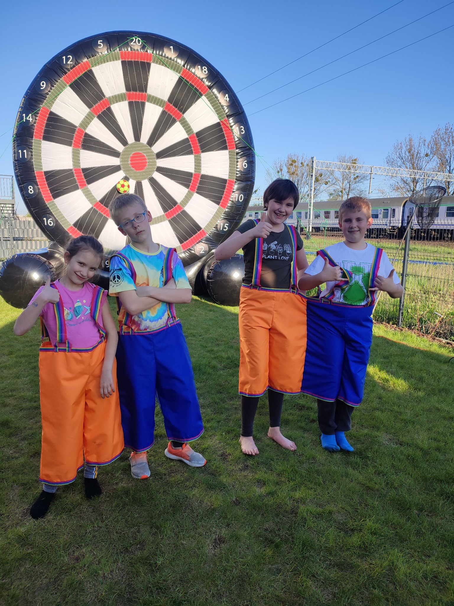 Grupa dzieci w kolorowych strojach prezentuje się na tle dmuchanej tarczy do darta, z pociągiem w tle, na zielonej trawie.