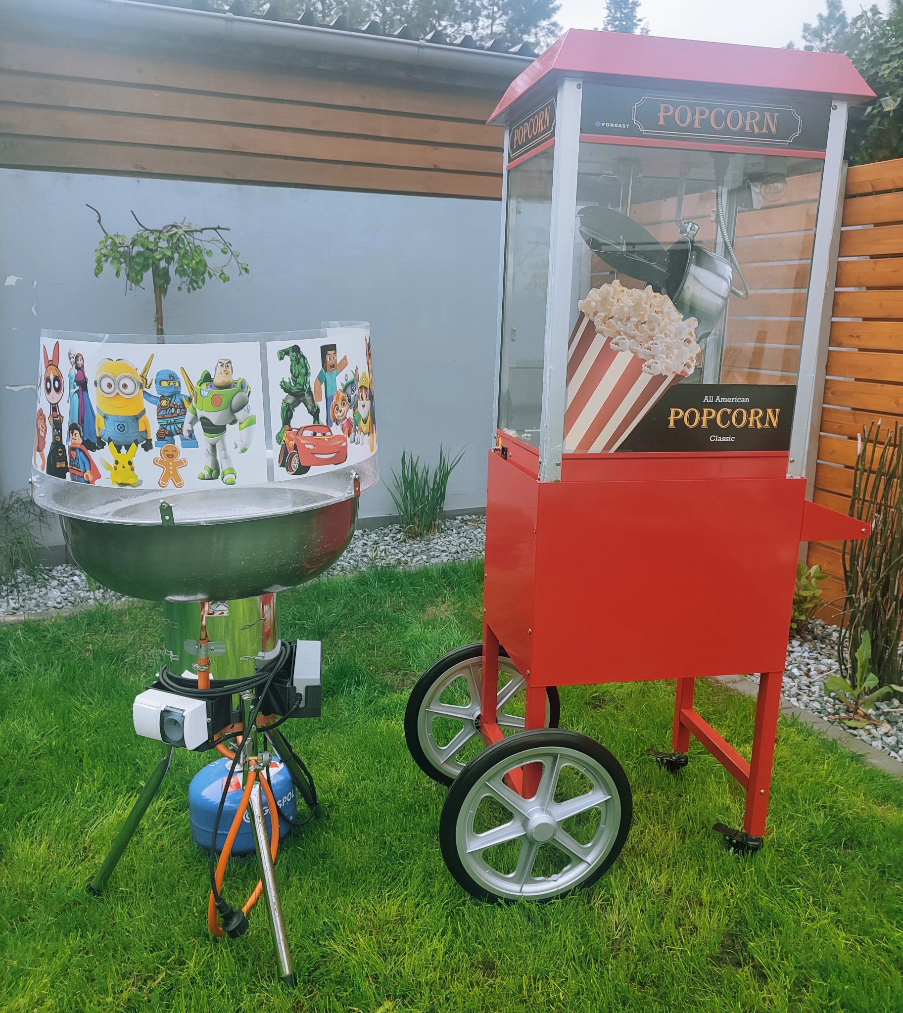Dwa urządzenia do przygotowywania i serwowania popcornu na trawie: profesjonalny wózek z popcornem w stylu retro z napisem 'Popcorn' oraz przenośny kociołek z osłoną z postaciami z bajek.