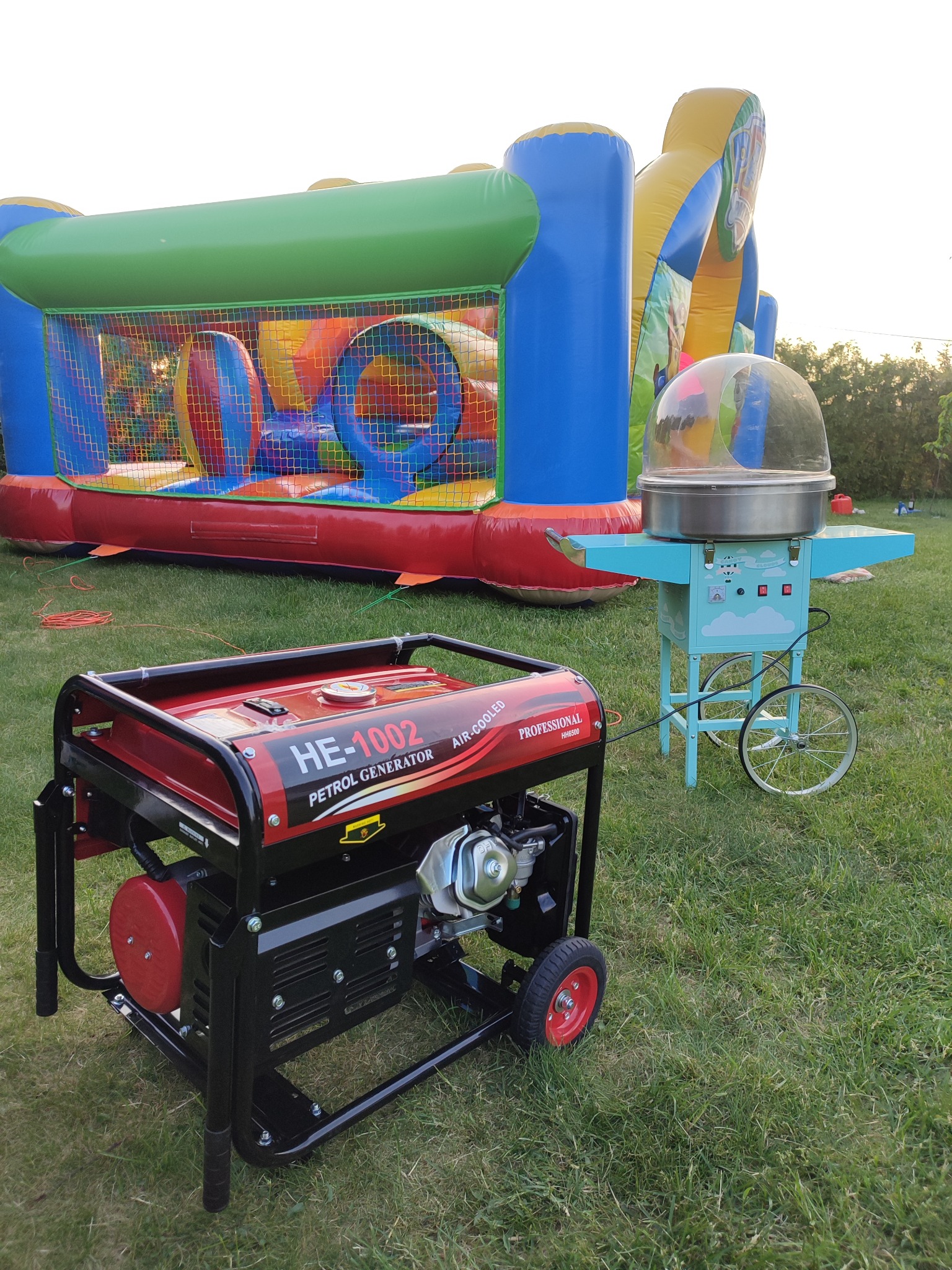 Rozłożony dmuchany plac zabaw z tunelem i zjeżdżalnią, maszyna do waty cukrowej na kółkach oraz czerwono-czarny generator spalinowy na trawie.