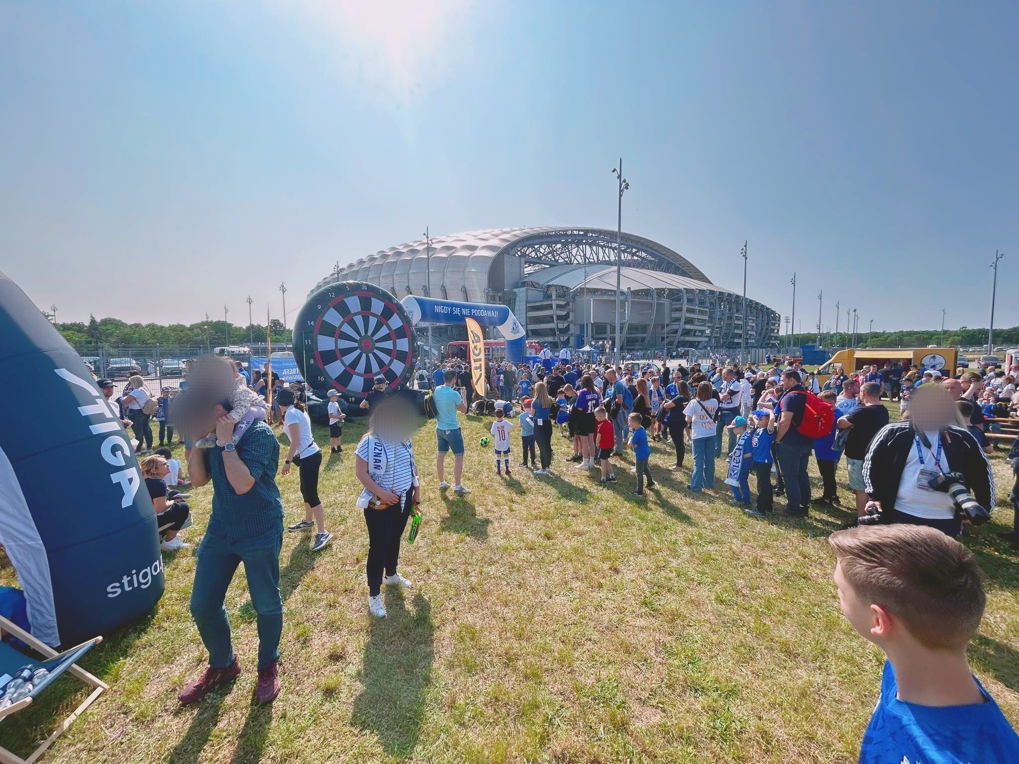Tłum ludzi na trawniku przed stadionem, dmuchana tarcza do darta i brama z napisem 'Nigdy się nie poddawaj'.