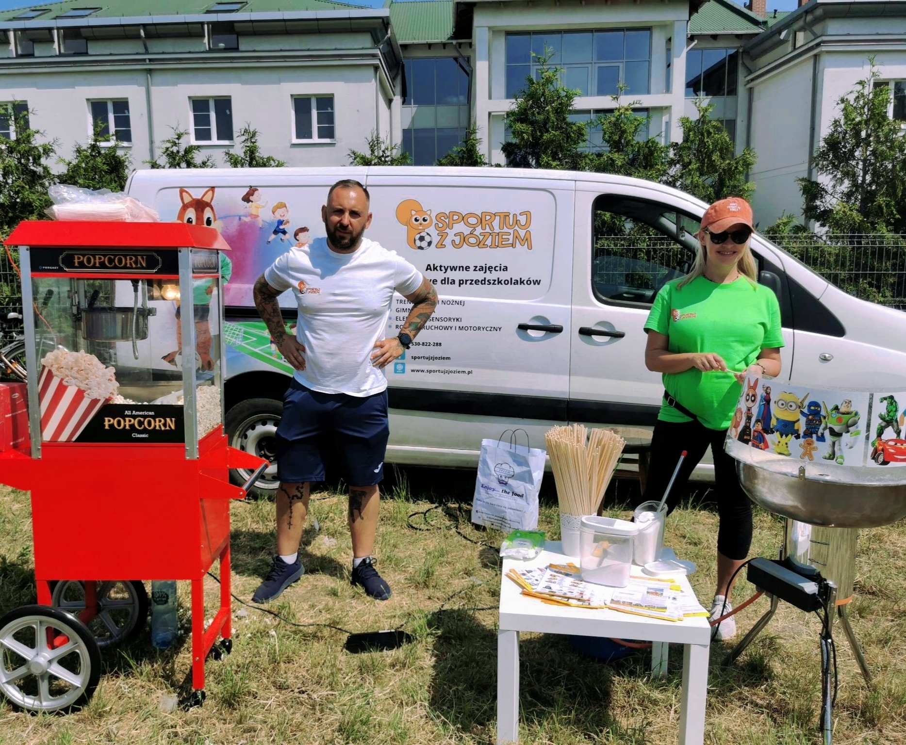 Dwóch animatorów na trawie obok wozu z popcornem i maszyny do waty cukrowej, na tle białego busa z logo firmy 'Sportuj z Józiem' oferującej zajęcia dla przedszkolaków.