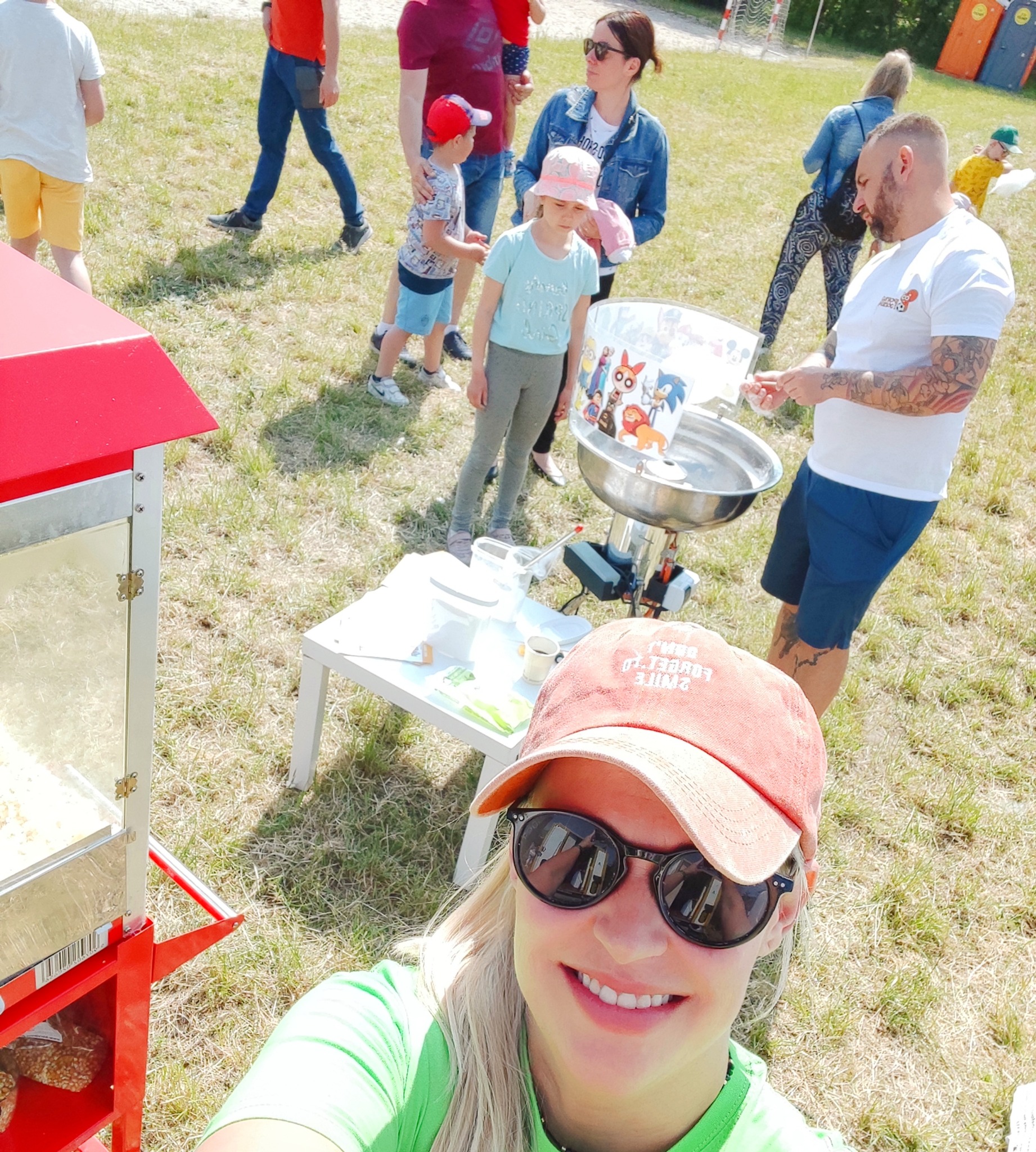 Selfie z operatorem maszyny do waty cukrowej w Poznaniu podczas imprezy plenerowej dla dzieci, z widoczną maszyną do waty, stolikiem i grupą dzieci oraz dorosłych w tle.