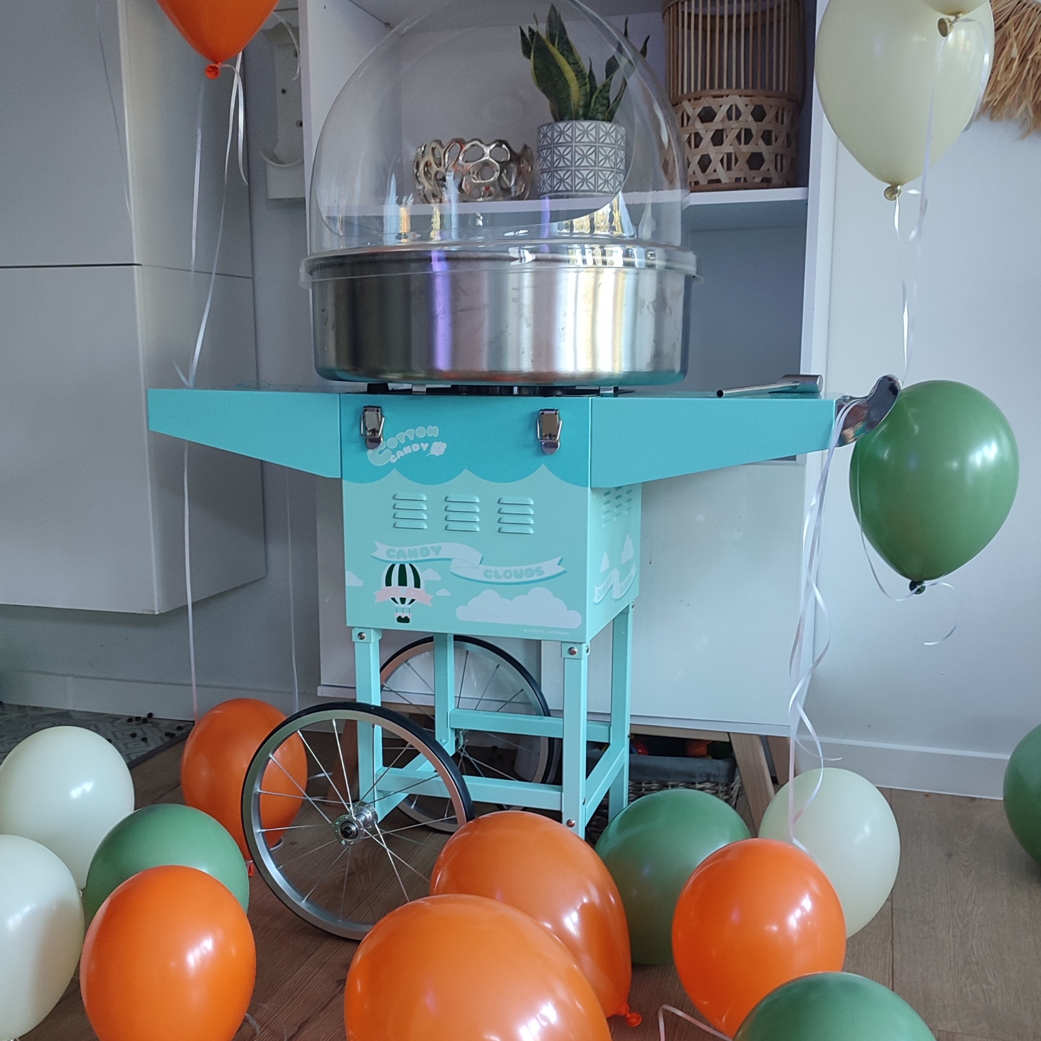 Turkusowa maszyna do waty cukrowej z napisem 'Cotton Candy Clouds' otoczona pomarańczowymi, zielonymi i białymi balonami w pomieszczeniu.