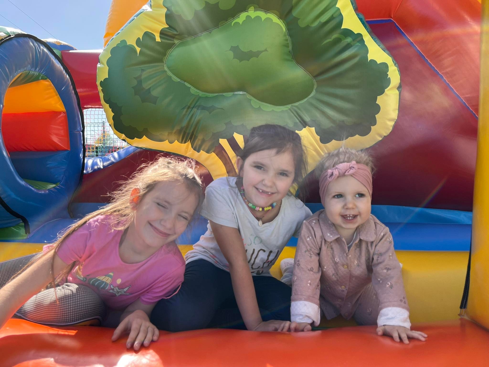 Trzy uśmiechnięte dziewczynki w różnym wieku siedzą w dmuchanym zamku z motywem drzewa, w tle widać siatkę zabezpieczającą.