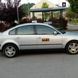 Srebrny samochód osobowy z oznaczeniami Taxi Mszana Dolna zaparkowany na tle przystanku autobusowego z napisem TAXI.