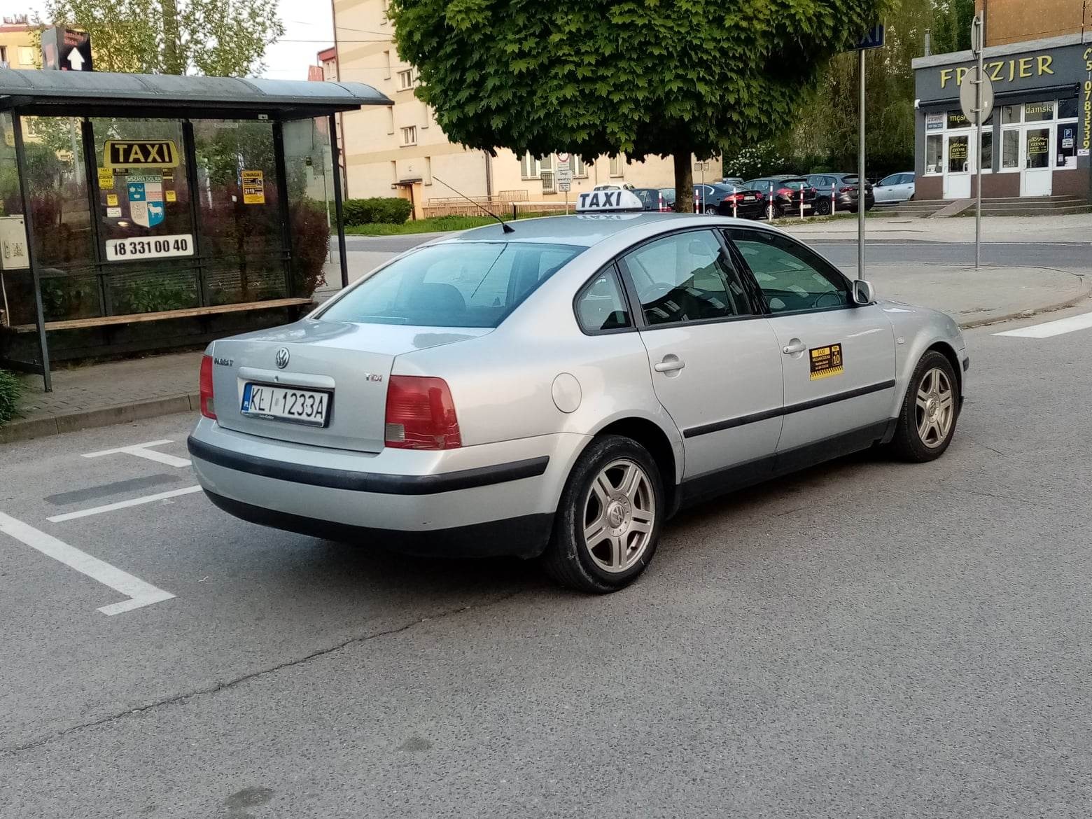 Srebrny Volkswagen Passat z oznaczeniami taxi, zaparkowany obok przystanku autobusowego z reklamą taxi w Mszanie Dolnej, widoczny budynek fryzjera w tle.