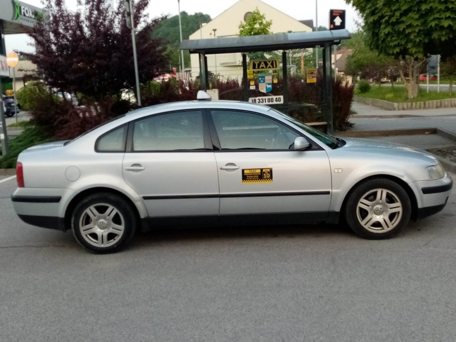 Srebrny samochód osobowy z oznaczeniami Taxi Mszana Dolna zaparkowany na tle przystanku autobusowego z napisem TAXI.