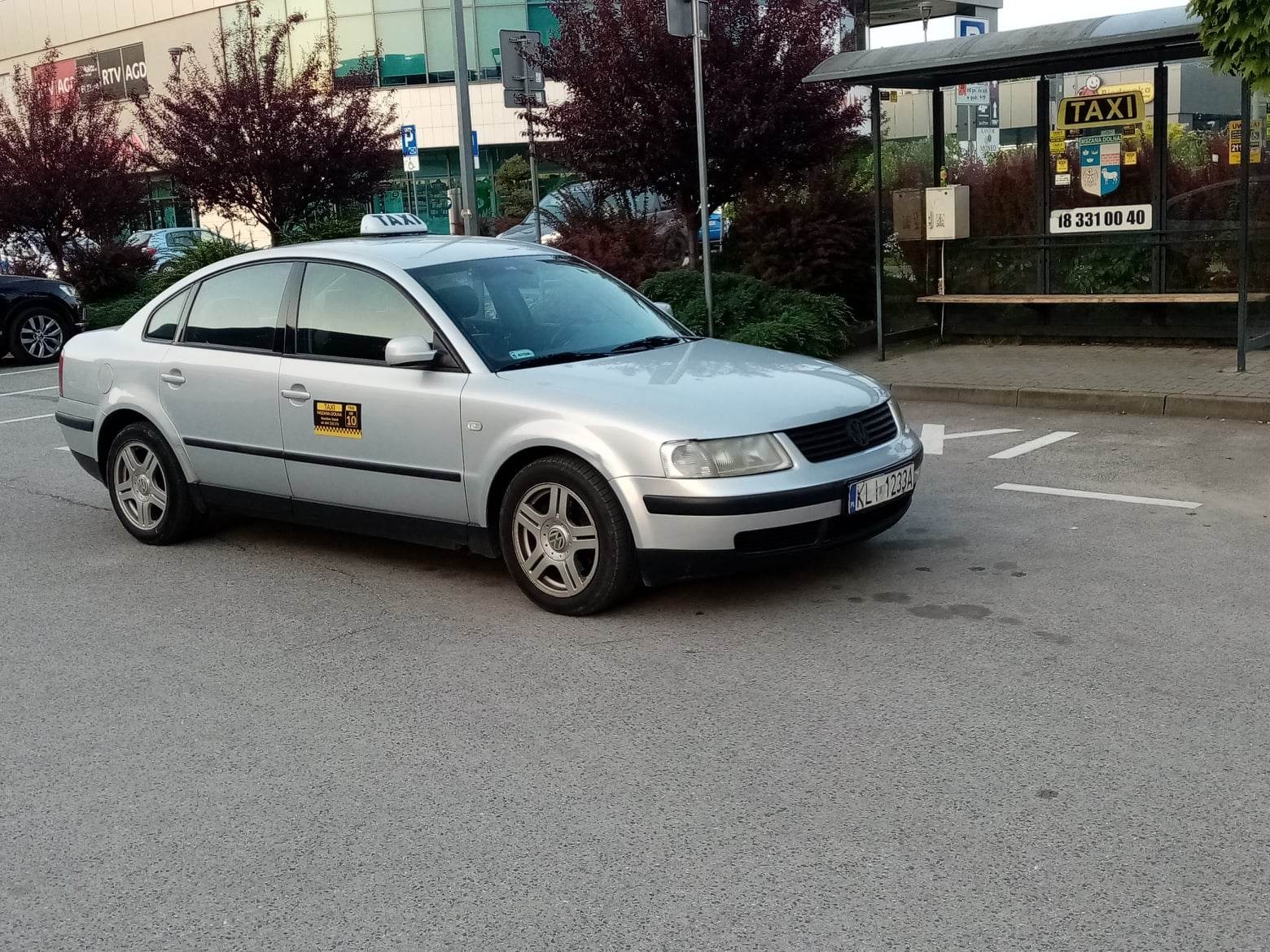 Srebrny samochód osobowy z napisem TAXI na dachu, zaparkowany obok przystanku autobusowego z reklamą taksówki w Mszanie Dolnej, widoczny numer rejestracyjny KL 12334.