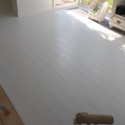 REMONT BG - Biała, świeżo polakierowana podłoga z paneli, z widoczną drabiną malarską na kafelkach i wałkiem malarskim w prawym dolnym rogu, w tle białe meble i okno z wpadającym światłem.