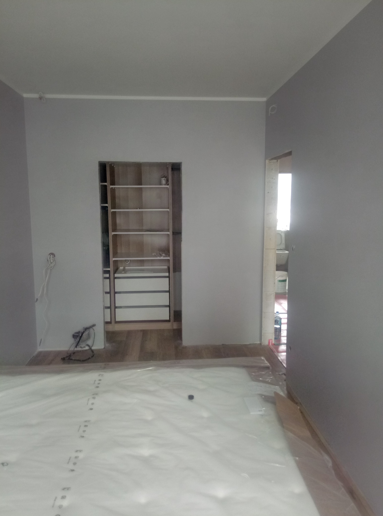 Szare ściany w pokoju przed wykończeniem, podłoga zabezpieczona folią, widoczna zabudowa szafy wnękowej z półkami i szufladami oraz wejście do łazienki z widoczną toaletą i wiadrem.