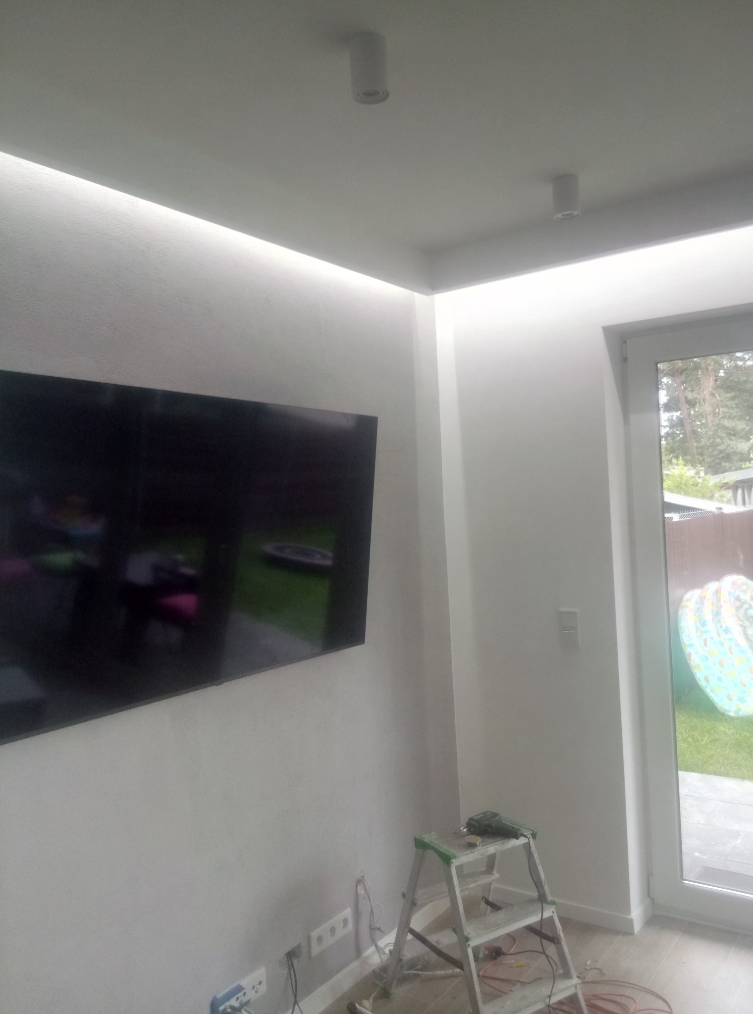 Białe wnętrze po remoncie, z podświetleniem LED na suficie, telewizorem na ścianie, drabinką i narzędziami, widok na ogród przez okno.