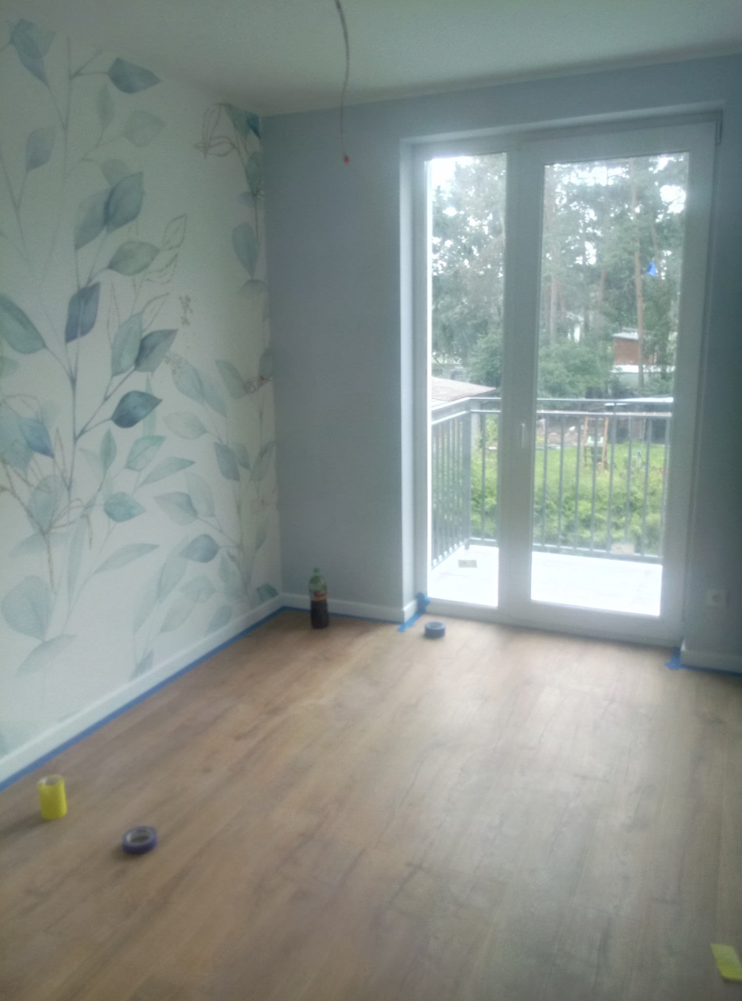 Pusty pokój z drewnianą podłogą i ścianą z motywem roślinnym, zabezpieczony taśmą malarską przy podłodze, z balkonem widocznym przez przeszklone drzwi balkonowe.