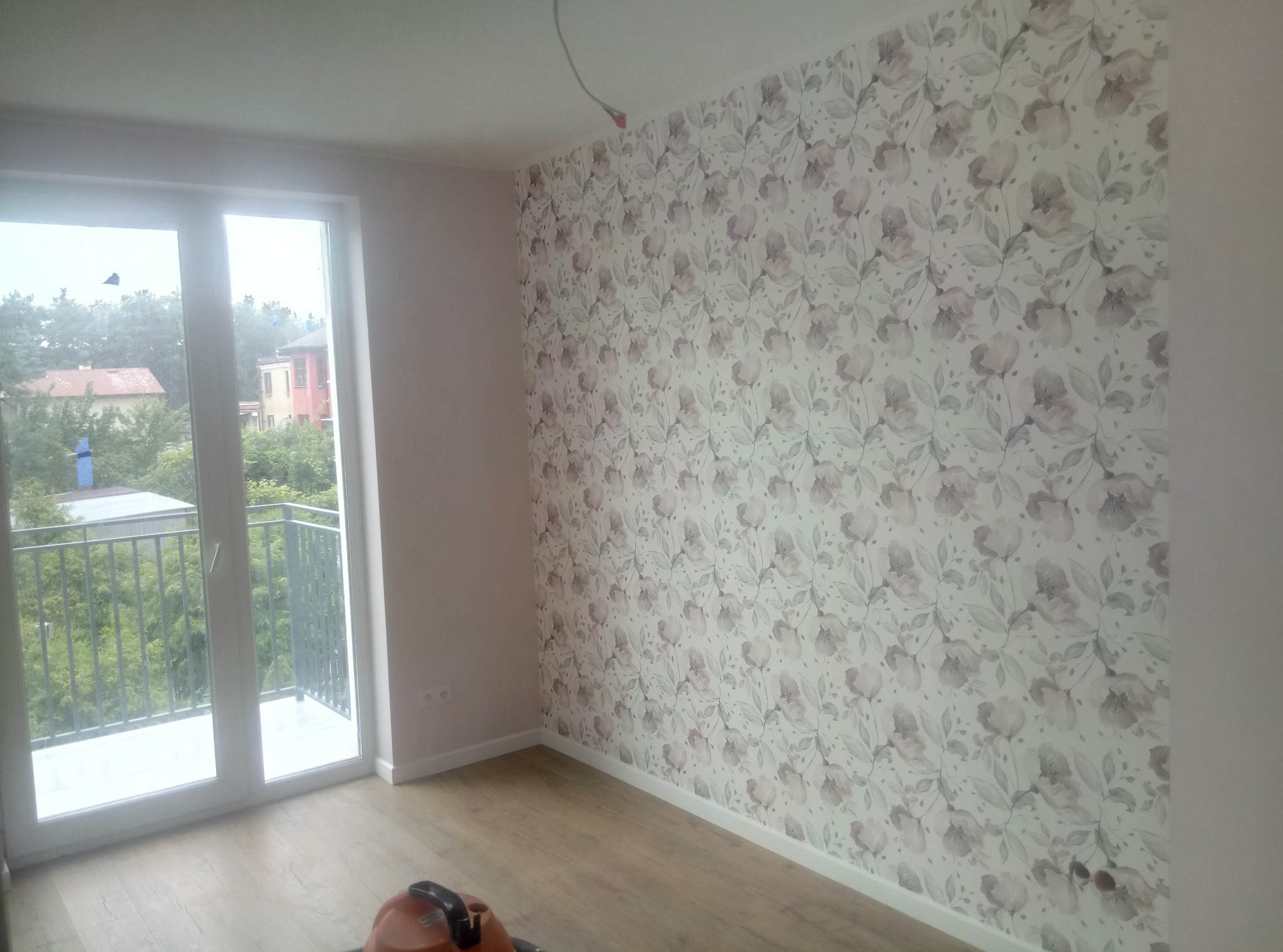 Pokój po remoncie: ściana pokryta tapetą w kwiaty, widok na balkon i zieleń za oknem, na podłodze odkurzacz.