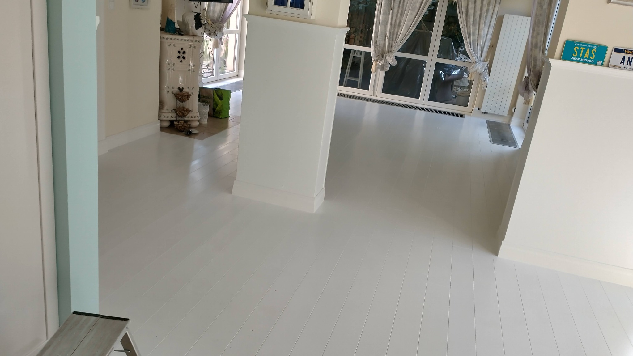 Jasne, białe deski podłogowe po renowacji, widok z perspektywy podłogi, fragment drabiny malarskiej w rogu, w tle okno z zasłonami i fragment dekoracyjnego pieca.