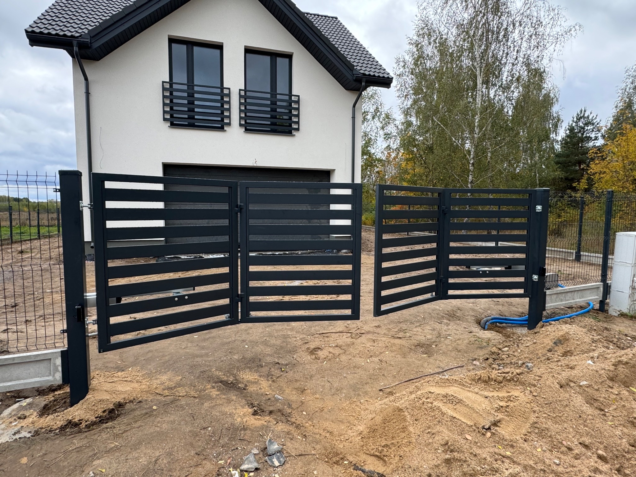 Nowoczesna, grafitowa brama wjazdowa z poziomymi panelami na tle jasnego domu z czarnym dachem i oknami. Częściowo otwarte skrzydła bramy.