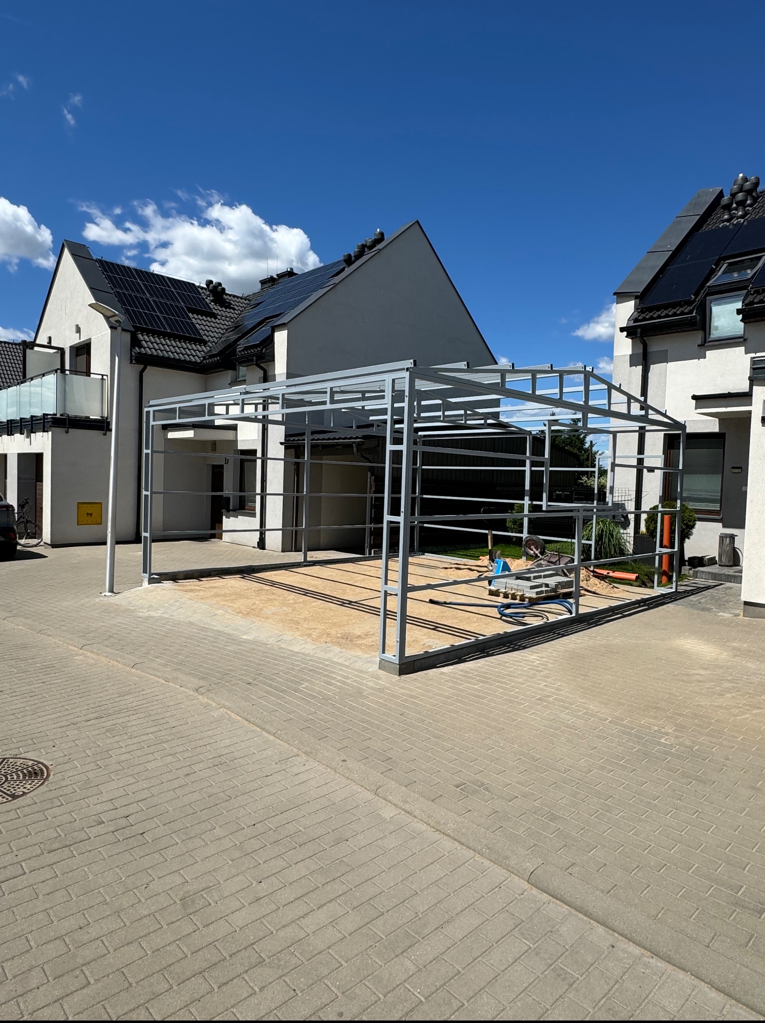 Metalowa konstrukcja w trakcie budowy w przestrzeni między domami. Widoczne narzędzia i materiały budowlane. Błękitne niebo z chmurami w tle.