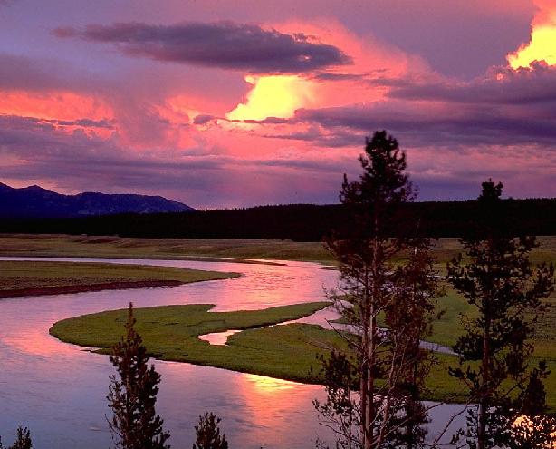 Malowniczy krajobraz rzeki wijącej się przez zielone tereny pod rozświetlonym różowo-fioletowym niebem o zachodzie słońca, z sylwetkami gór i lasów w tle.