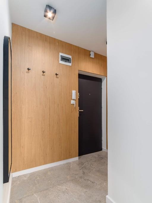 Nowoczesny przedpokój z drewnianą zabudową ścienną, wbudowanymi haczykami i czarnymi drzwiami wejściowymi, oświetlony punktowo, z podłogą z płytek imitujących marmur.