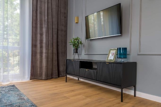 Nowoczesny salon z szarymi ścianami, telewizorem na ścianie, czarną szafką RTV z dekoracjami, brązowymi zasłonami i drewnianą podłogą.