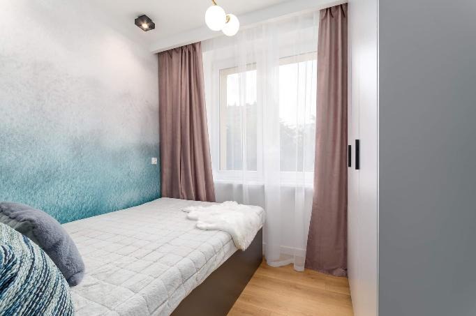 Nowoczesna sypialnia z ombre na ścianie w odcieniach bieli i błękitu, jasnym łóżkiem, brązowymi zasłonami i białymi firankami w oknie oraz minimalistyczną lampą sufitową.