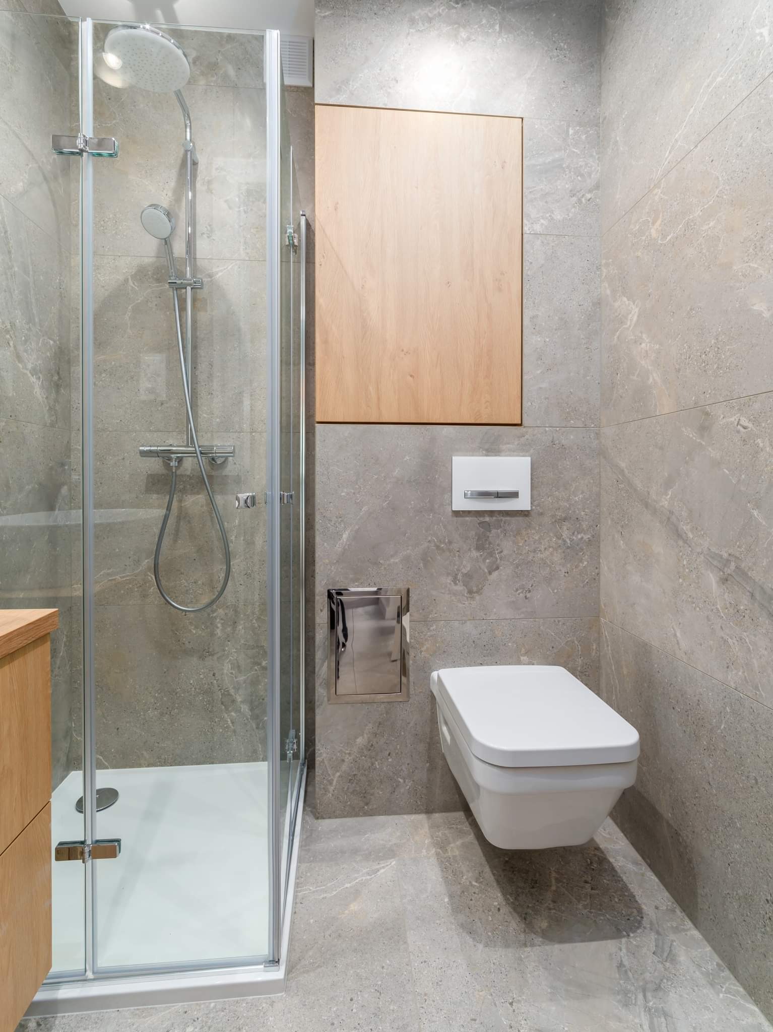 Nowoczesna łazienka z szarymi płytkami imitującymi marmur, kabiną prysznicową ze szklanymi drzwiami i białym brodzikiem, podwieszaną miską WC oraz szafką z drewnianym frontem