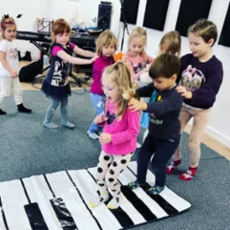 Grupa dzieci bawi się na macie imitującej klawiaturę fortepianu w jasnym pomieszczeniu z pianinem i panelami akustycznymi na ścianie.